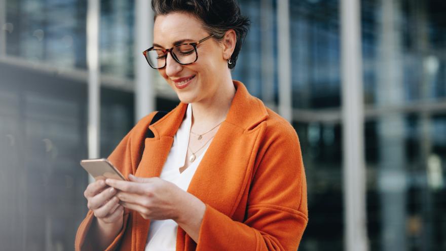 Eine Frau mittleren Alters mit Brille hält ihr Smartphone in den Händen, blickt darauf und lächelt mit offenem Mund. Sie trägt einen ockerfarbenen Mantel, hinter ihr ist ein verglastes Gebäude.