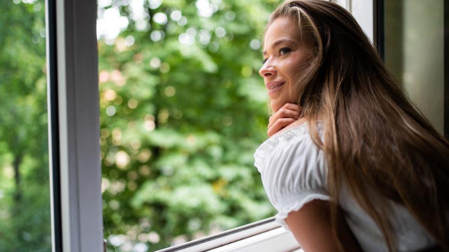Eine junge Frau lehnt sich auf die Fensterbank eines offenen Fensters und blickt nach draußen ins Grüne. 