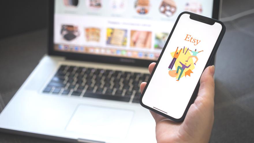 Eine Hand hält ein Handy, zu sehen die Etsy-App, im Hintergrund ein Laptop mit dem Etsy-Online Shop
