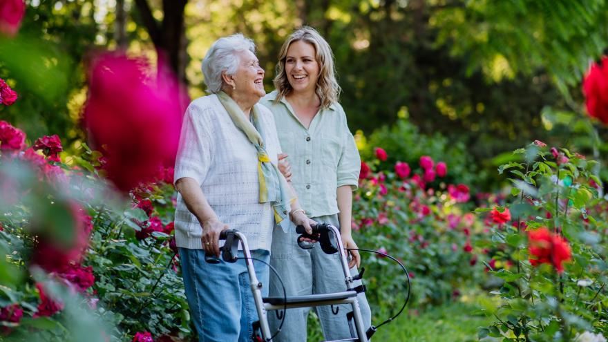 Eine alte Frau mit Rollator und eine junge Frau sind zusammen in einem von roten Rosen übersäten Park. Die junge Frau stützt den Arm der alten Frau. Beide lachen und sehen glücklich aus.
