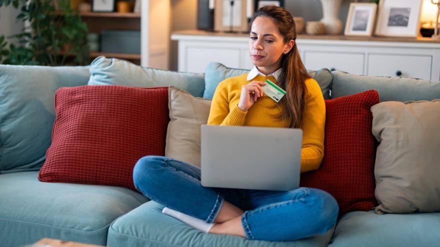 Eine Frau sitzt im Schneidersitz auf dem Sofa. Sie hat einen Laptop auf dem Schoß und eine Kreditkarte in der Hand und schaut prüfend.