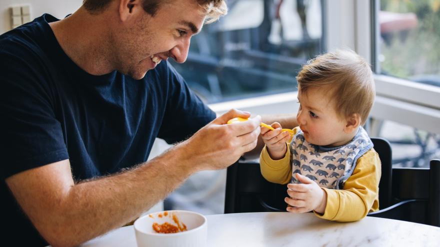 Ein Mann mittleren Alters sitzt mit einem Baby am Tisch und füttert es.