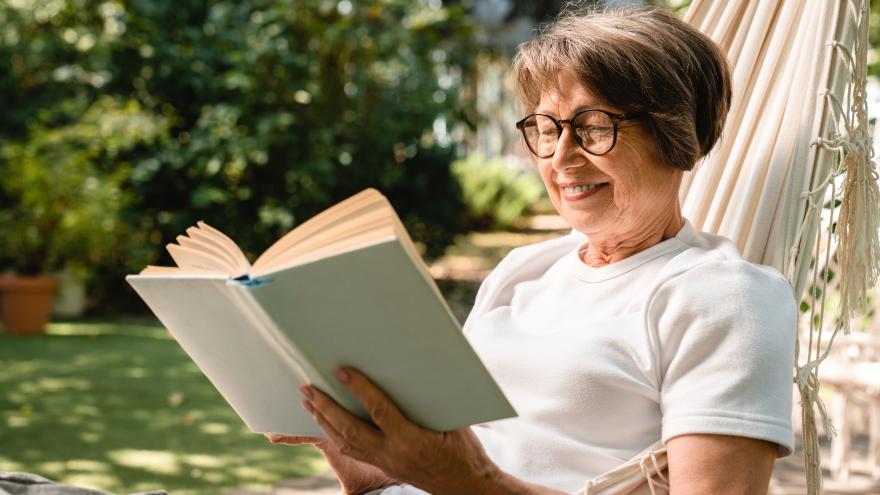 Eine alte Frau liegt entspannt im Garten auf einer Hängematte, liest ein Buch und lächelt zufrieden.