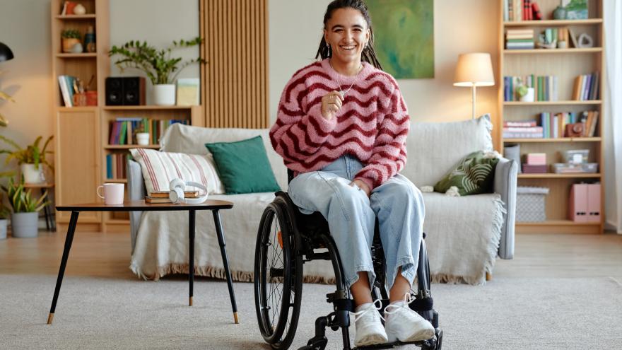 Eine junge Frau sitzt im Rollstuhl im Wohnzimmer vor dem Sofa. Sie lacht in die Kamera.