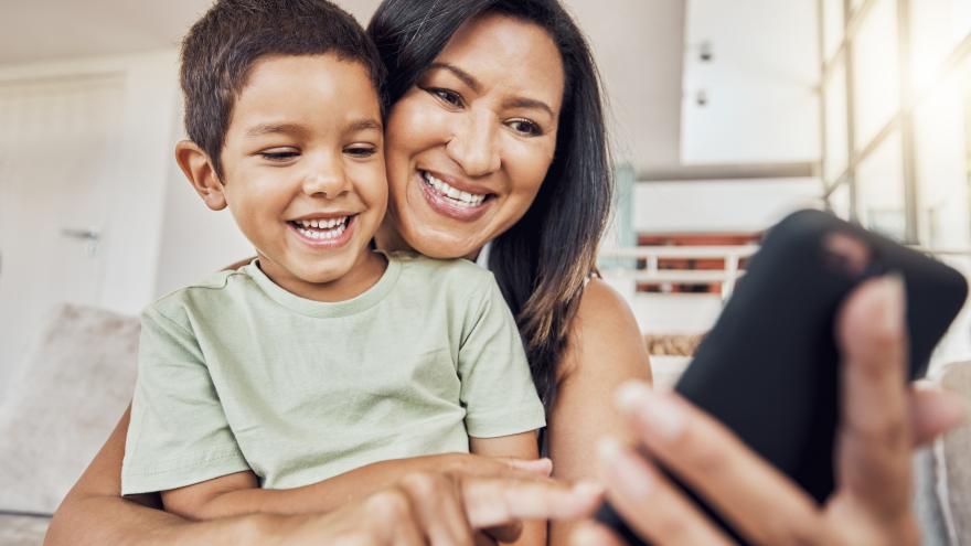 Eine Frau hat einen kleinen Jungen im Arm. In ihrer Hand hält sie ein Smartphone. Beide lachen und schauen auf das Handy.