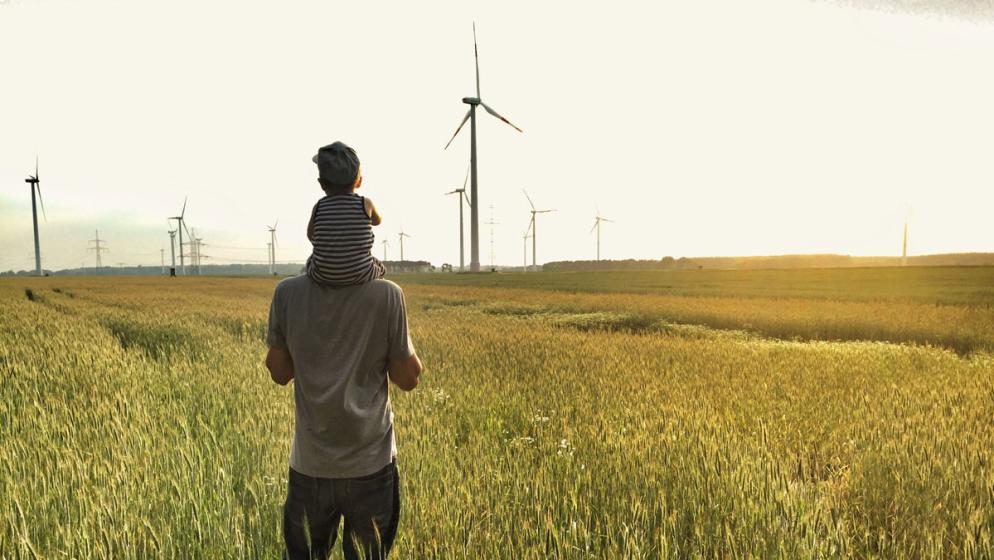 Vater trägt Kind auf den Schultern auf einem Feld vor Windrädern bei Sonnenuntergang.