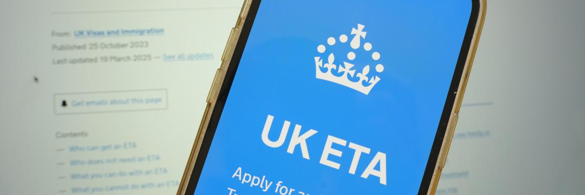 Ein Smartphone zeigt die UK-Regierungswebsite zur Beantragung der elektronischen Reisegenehmigung (ETA) für das Vereinigte Königreich an. Im Hintergrund ist ein verschwommener Bildschirm mit weiteren Informationen zur ETA sichtbar.