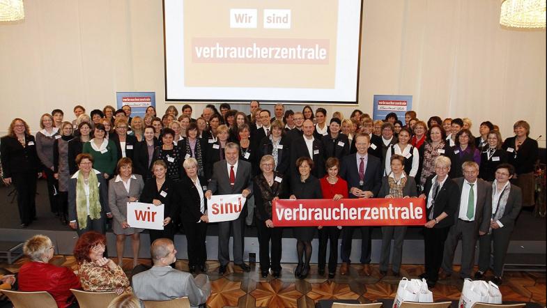 Viele Menschen auf eienr Bühne, die in die Kamera blicken und ein Schild vor sich halten.