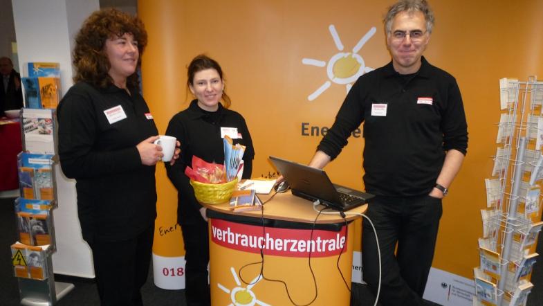 Drei Menschen stehen an einem Informationsstand und lachen in die Kamera.