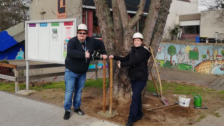 Zwei Personen mit Bauarbeietrhelm udn Werkzeug an einem Baum.