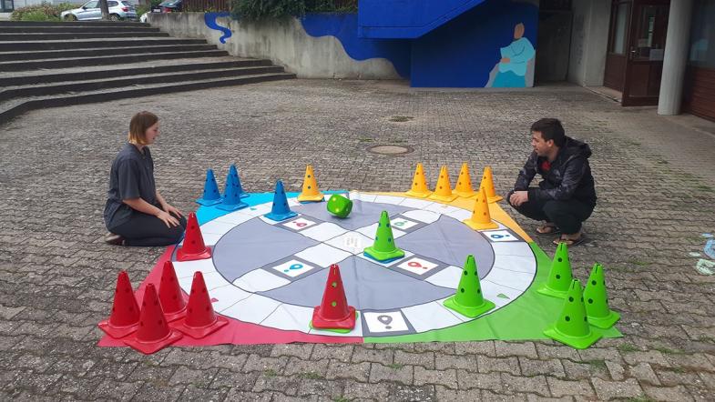 Zwei Erwachsene sitzen auf dem Bürgersteig vor einem sehr großen Spiel mit Spielmännchen.
