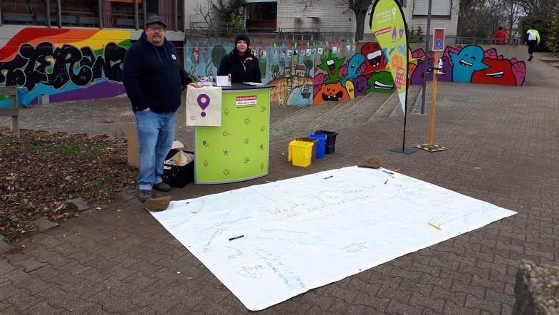 Zwei Menschen stehen vor einem Infostand und blicken in die Kamera.