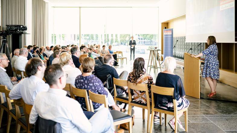 Eine Frau steht am Rednerpult und links von ihr sieht man das Publikum im Saal.