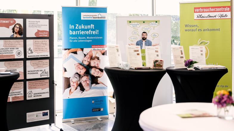 Ein Messestand mit zwei Stehtischen und Plakaten im Hintergrund.