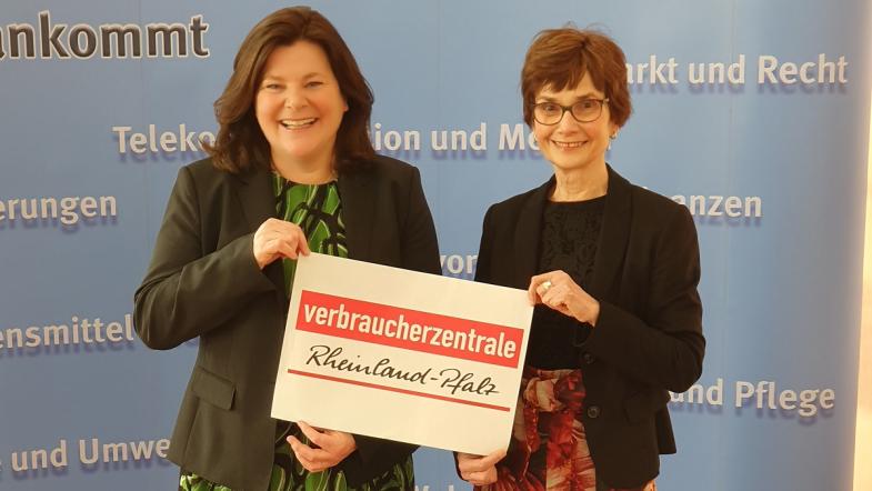 Zwei Frauen stehen vor einer baluen Wand und halten ein Schild mit Verbraucherzentrale Rheinland-Pfalz zwischen sich.