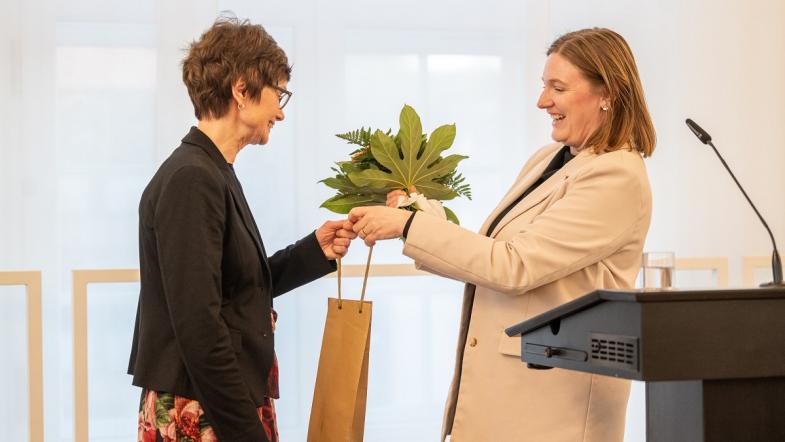 Rechts steht eine Frau an einem Rednerpult und überreicht einer anderen Frau links im Bild einen Blumenstrauß und eine Geschenktüte.