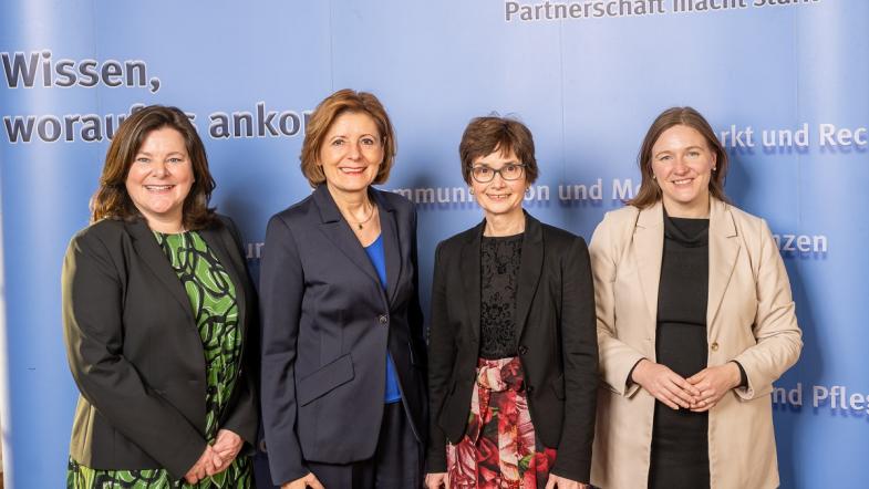 Vier Frauen stehen vor einer blauen Wand und blicken in die Kamera.