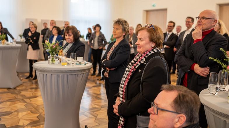 Einige Menschen stehen an Stehtischen vor einer Bühne und verfolgen das Geschehen auf der Bühne.