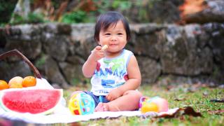 Ein Kleinkind sitzt auf einer Picknickdecke und isst ein kleines Stück Obst. Links neben ihm steht ein großes Stück Wassermelone.