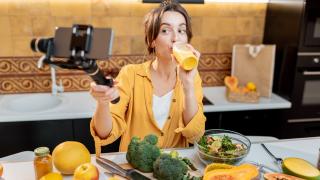 Frau mit Smoothie ind er einen Hand udn Smartphone in der anderen. Sie filmt sich beim Trinken. Vor ihr ist ein Schneidebrett und einiges an Obst und Gemüse