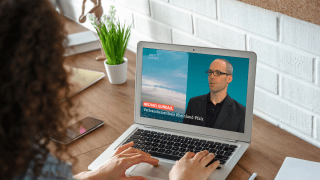 Eine Frau an einem Laptop. Auf dem Bildschirm ist ein Mann zu sehen, der gerade etwas erklärt.