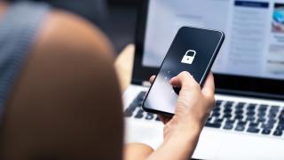 Eine Person sitzt an einem Laptop und hält ein Smartphone in der Hand. Auf dem Smartphone ist ein Verschlüsselungssymbol.