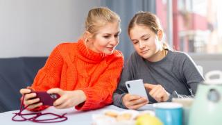 Mutter sitzt mit Tochter am Tisch und schaut auf ihr Smartphone.