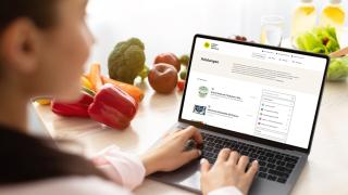 Eine Frau sitzt mit Laptop am Küchentisch und schaut auf eine Internetseite auf dem Bildschirm.