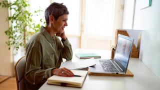 Frau sitzt am Tisch und schaut lächelnd auf den Bildschirm ihres Laptops.