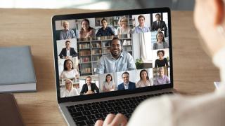 Jemand sitzt vor einem Laptop und hält eine digitale Konferenz ab. Auf dem Bildschirm sind viele Menschen zu sehen, die in die KAmera schauen. 