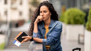Eine Frau steht mit ihren Reiseunterlagen in der Hand auf der Straße. Sie sieht besorgt aus.