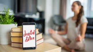 Im Vordergrund liegt ein Handy mit den Wörtern: digital detox auf dem Bildschirm. Im Hintergrund macht eine Frau Yoga