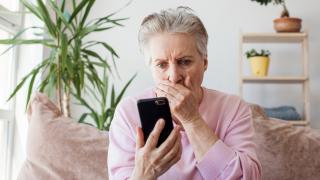 Eine ältere Frau sitzt auf dem Sofa und ist schockiert über den Anruf, den sie gerade erhalten hat.
