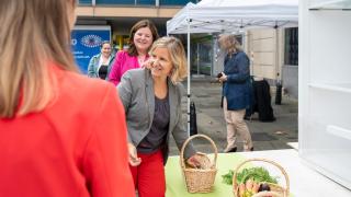 Zwei Frauen stehen an einem Tisch und blicken nach hinten. Eien Frau greift dabei in einen Korb vor ihr.