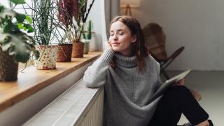 Eine Frau sitzt mit geschlossenen Augen und entspannt vor einer Heizung in einem Zimmer. Sie hält ein Tablet in der Hand und neben ihr auf der Fensterbank stehen verschiedene Zimmerpflanzen.