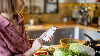 Frau sitzt am Küchentisch mit Lebensmittel und einem Smartphone.