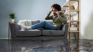 Ein Mann sitzt auf seiner Couch und hat ein Handy am Ohr. Seine Wohnung steht voller Wasser.