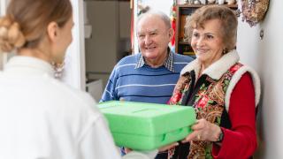 Eine junge Frau eines Lieferdienstes bringt zwei Senioren Essen in einer grünen Thermobox.