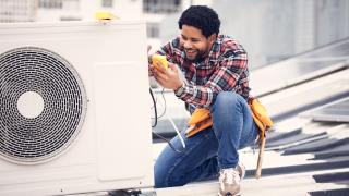 Ein freundlicher Techniker sitzt mit einem Testgerät neben einer elektrischen Wärmepumpe.