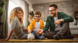 Eltern sitzen mit ihrem Kind auf dem Boden. Das Kind wirft Geldmünzen in ein Sparschwein.