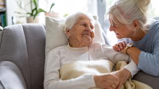Eine Seniorin Sitzt durch ein Kissen gestützt auf der Couch und lächelt. Eine weitere Frau lehnt sich liebevoll zu ihr rüber und schaut sie an.