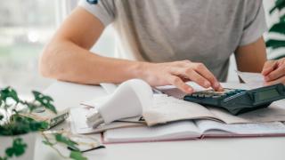 Ausschnitt eines menschlichen Torsos. Ein Mensch tippt auf einem Taschenrechner. Auf dem Tisch liegen außerdem eine LED-Birne und ein Schreibblock.