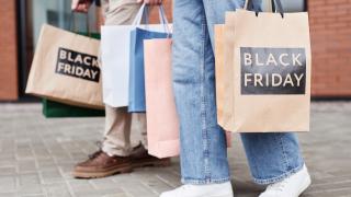 Menschen mit Papiertüten, auf denen die Worte Black Friday stehen, schlendern auf einer Straße. Es sind nur die Unterkörper zu sehen.