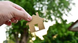 Eine Hand hält eine aus Pappe ausgeschnittenes Haus vor einen Baum