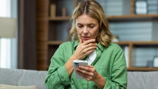 Eine Frau schaut fragend/ erschrockend auf ein Smartphone in ihrer Hand. Die andere Hand hält sie an ihr Kinn. 