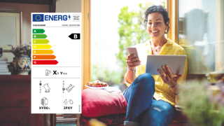 Frau am Fenster mit Tablet und Smartphone in der Hand, links im Bild das Energielabel für Smartphones und Tablets