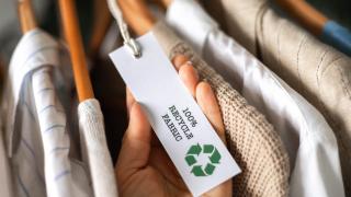 Kleidung auf Kleiderbügel, Hand hält Etikett auf dem steht "100% Recycle Fabric"