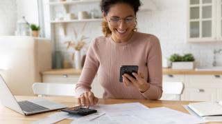 Frau sitzt am Schreibtisch mit vielen Rechnungen, sie schaut auf ihr Handy und tippt in den Taschenrechner