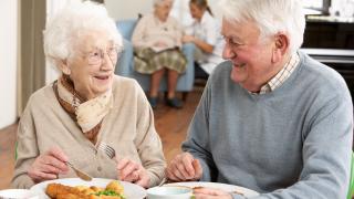 Alte Frau und alter Mann sitzen im Altenheim am Tisch, essen gemeinsam und lächeln sich an