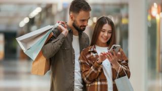 Mann und Frau sind in einer Einkaufspassage. Sie tragen Einkaufstüten und schauen gemeinsam auf ein Handy, das die Frau in ihrer Hand hält. Die Frau lacht, der Mann schaut zufrieden.